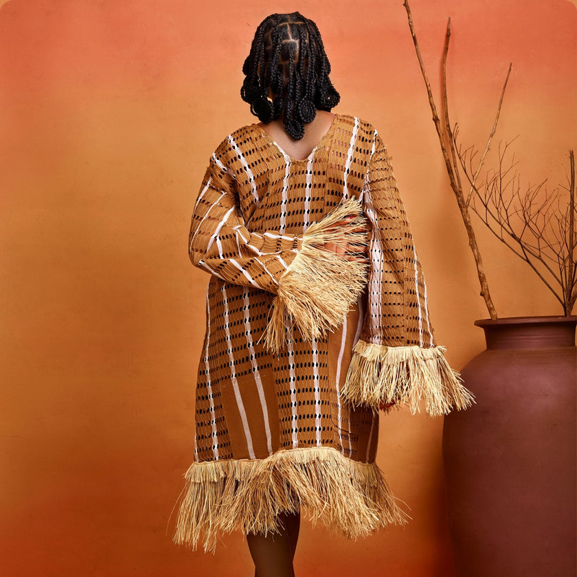 Back view : Rear view of the Asàkè Aso Oke dress showing deep V-neckline at the back, raffia-trimmed hem, and sleeve details as the model poses with one hand by her side and the other on her back.