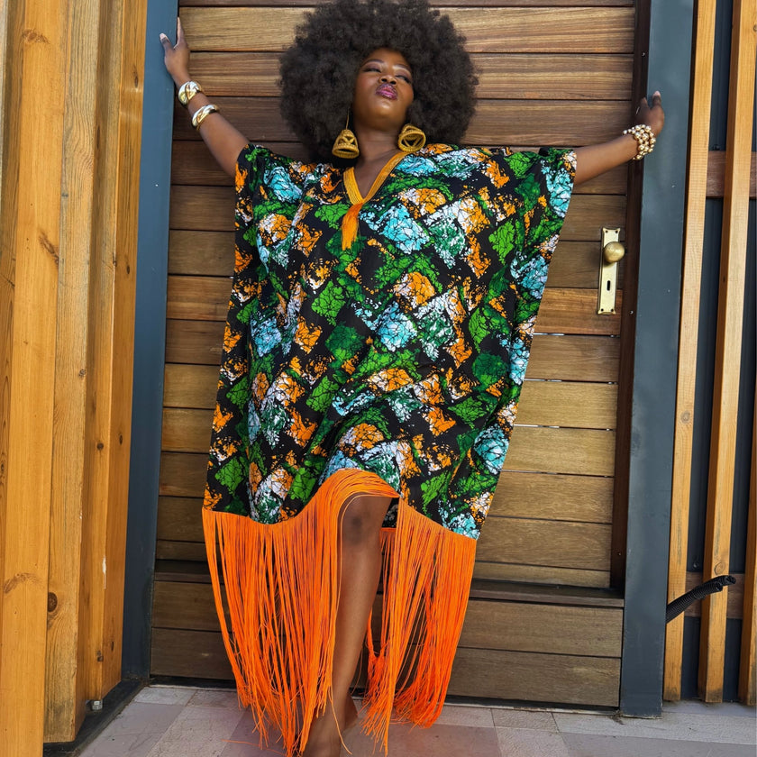 Front view with arms spread out : Model standing with hands spread wide to highlight the oversized flowing silhouette of the Jọkẹ́ Ankara bubu dress, showing its full width, lightweight fabric drape, and statement orange tassel hemline.