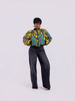 The front view of a cropped bomber jacket with a bold yellow and teal sunflower motif, with both hands inside the front pockets, styled with high-waisted black jeans.