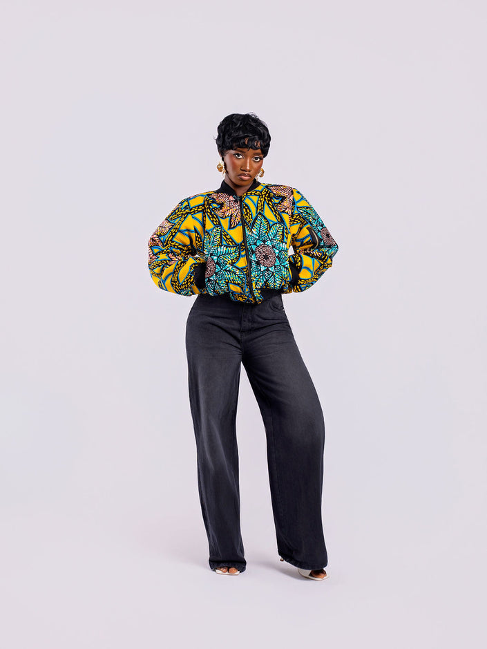 The front view of a cropped bomber jacket with a bold yellow and teal sunflower motif, with both hands inside the front pockets, styled with high-waisted black jeans.