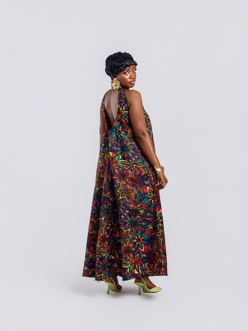 Full-length back view of a woman wearing the Amazin Apparels Kaleidoscope Gown, showing a large circular keyhole cutout and hidden elastic waistband in a vibrant floral African print.