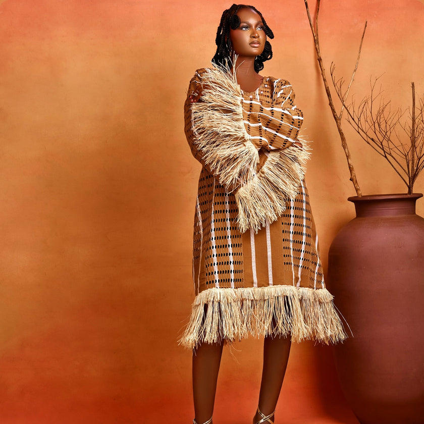 Full front view with crossed hands : Model in full front view with both hands crossed, highlighting the wide flowing sleeves, raffia fringe trims, and hemline of the handwoven Asàkè heritage maxi dress.