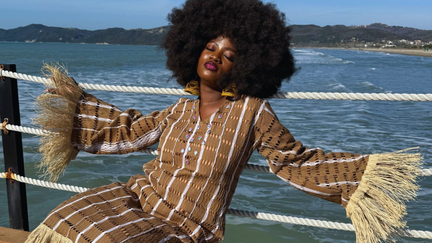 Model wearing Amazin Apparels’ Asake Aso-oke dress, sitting confidently by the ocean. Afrocentric elegance and freedom.
