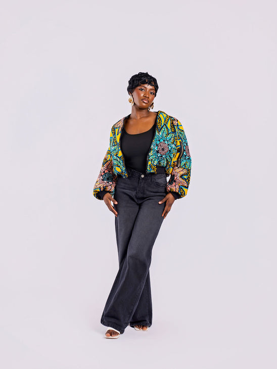 The front view of a cropped bomber jacket with a bold yellow and teal sunflower motif, with the front zip open, styled with high-waisted black jeans.