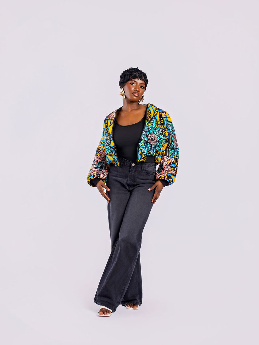 The front view of a cropped bomber jacket with a bold yellow and teal sunflower motif, with the front zip open, styled with high-waisted black jeans.