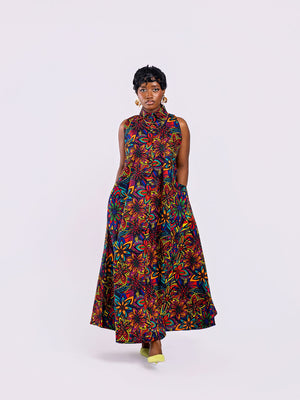 Full-length front view of a woman wearing the Amazin Apparels Kaleidoscope Gown, showing both hands tucked into the hidden side-seam pocket of the Kaleidoscope floral maxi dress, highlighting functional design. in a vibrant floral African print.