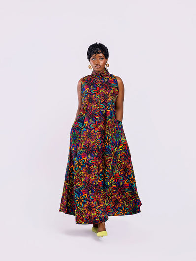 Full-length front view of a woman wearing the Amazin Apparels Kaleidoscope Gown, showing both hands tucked into the hidden side-seam pocket of the Kaleidoscope floral maxi dress, highlighting functional design. in a vibrant floral African print.