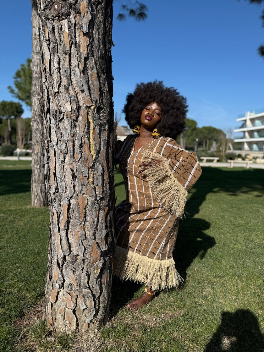 Side view : Side profile of the Asàkè maxi dress showcasing raffia fringe details on the sleeve and hemline, handwoven textured fabric, and structured yet fluid movement.