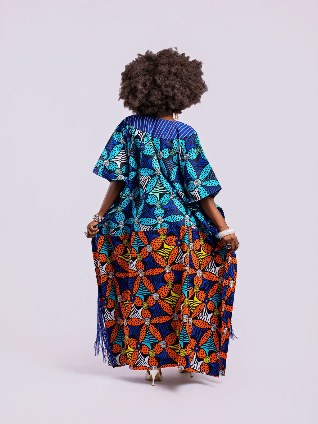 back view of a woman in the Amazin Apparels Aso Oke Fusion Gown, featuring a blue pinstripe top, bold floral skirt, and floor-length shredded Aso Oke fringe on the sides.