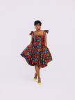 front view of a model wearing a straight neckline floral gown with a small V-cut and stylish folded details and structured bell-shaped skirt by Amazin Apparels, featuring red, blue, and green flowers on a black background.