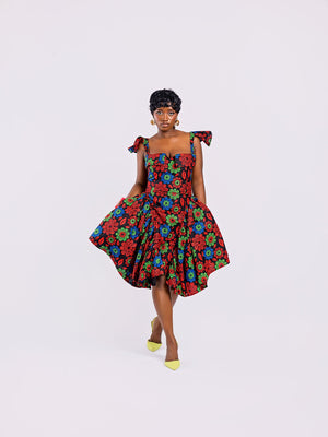 front view of a model wearing a straight neckline floral gown with a small V-cut and stylish folded details and structured bell-shaped skirt by Amazin Apparels, featuring red, blue, and green flowers on a black background.
