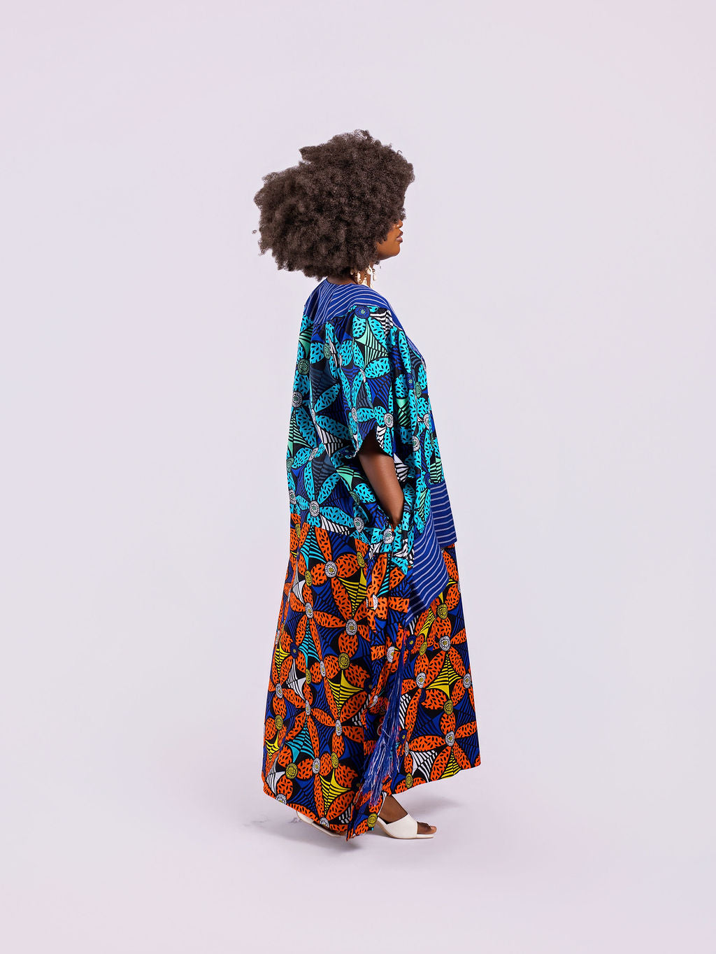A side view showing the dramatic movement of the blue shredded Aso Oke fringe and the high side slit of the Fusion Gown and A hand tucked into the hidden side-seam pocket, highlighting functional design.